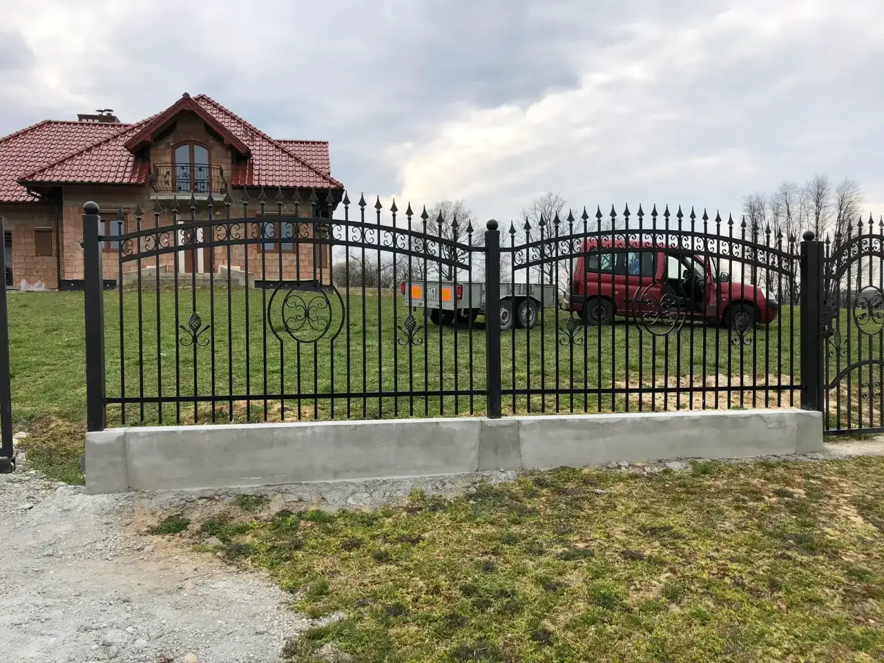Nowoczesne płoty kute z ozdobnymi elementami, otaczające posesję z domem i czerwonym samochodem.