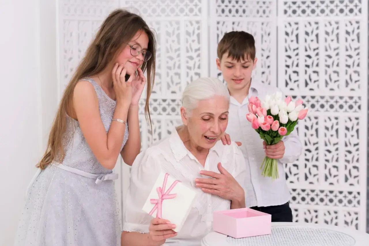 Wzruszona babcia otwiera prezent na Dzień Babci i Dziadka. Dzieci wręczają jej bukiet tulipanów i pudełko z upominkiem.