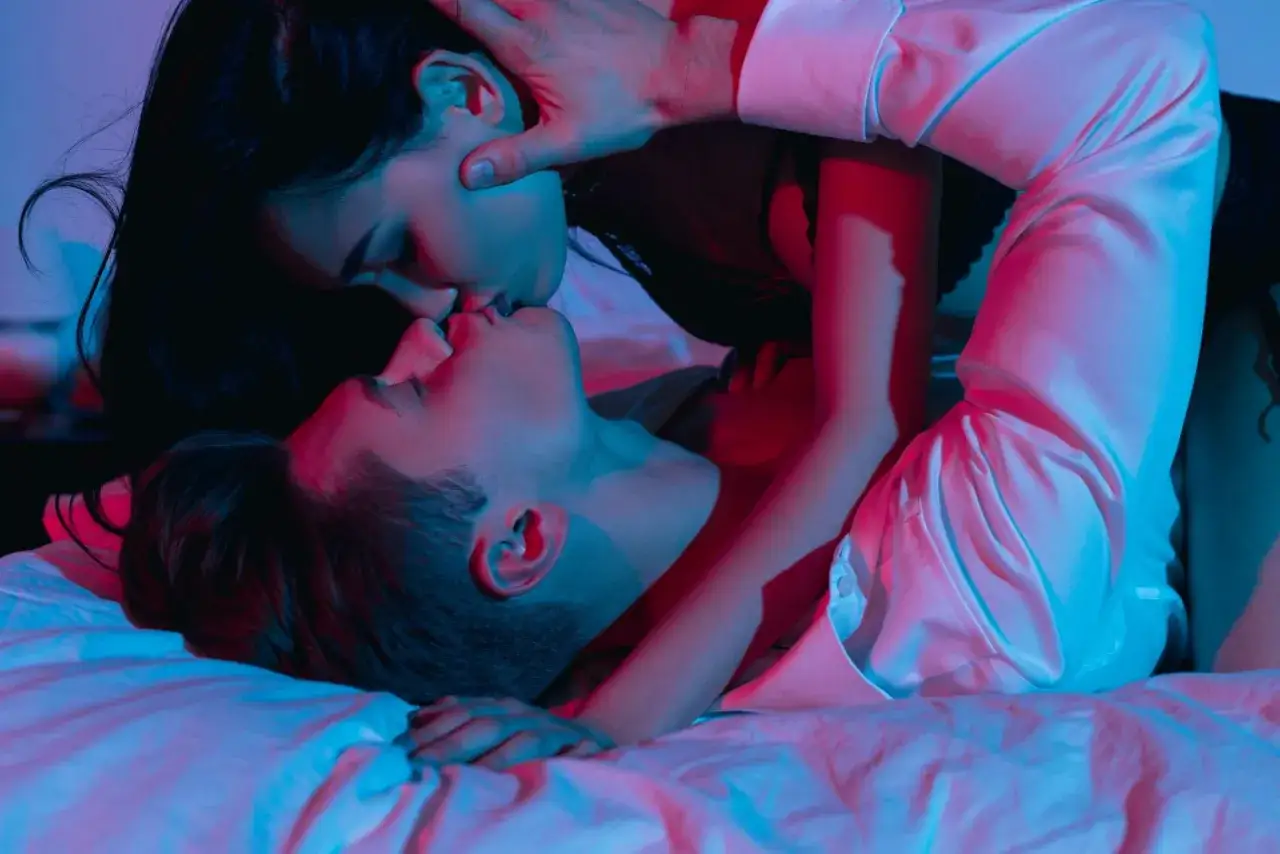 Para zbliża się do siebie w intymnym uścisku, tworząc ciekawe pozycje seksualne. Czerwone i niebieskie światła podkreślają ich bliskość.
