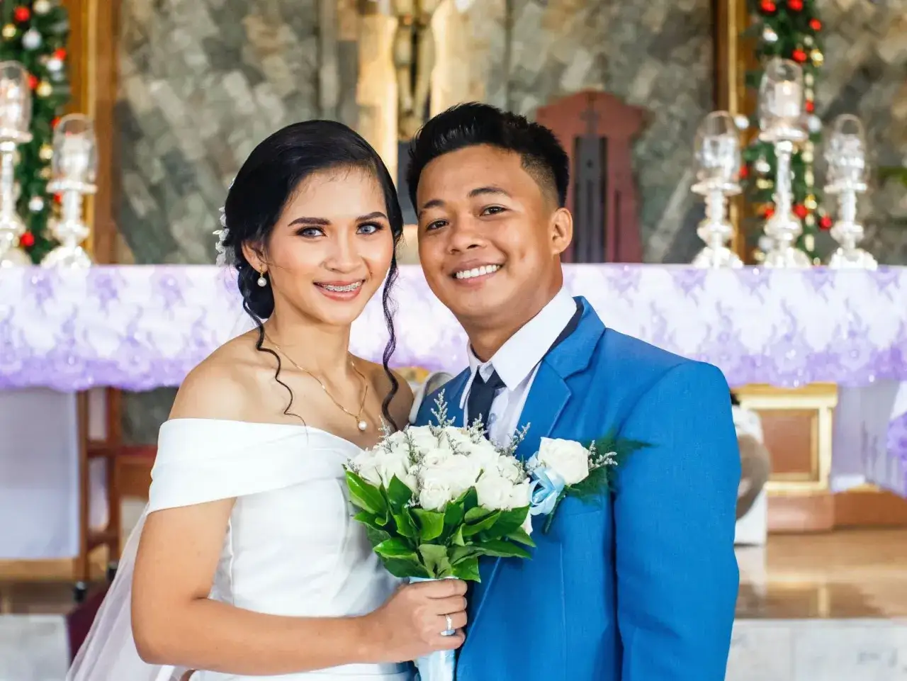 Novios sonriendo, ella con vestido blanco y él con traje azul. Un ramo de rosas blancas. Agradecimiento a los novios por la boda.