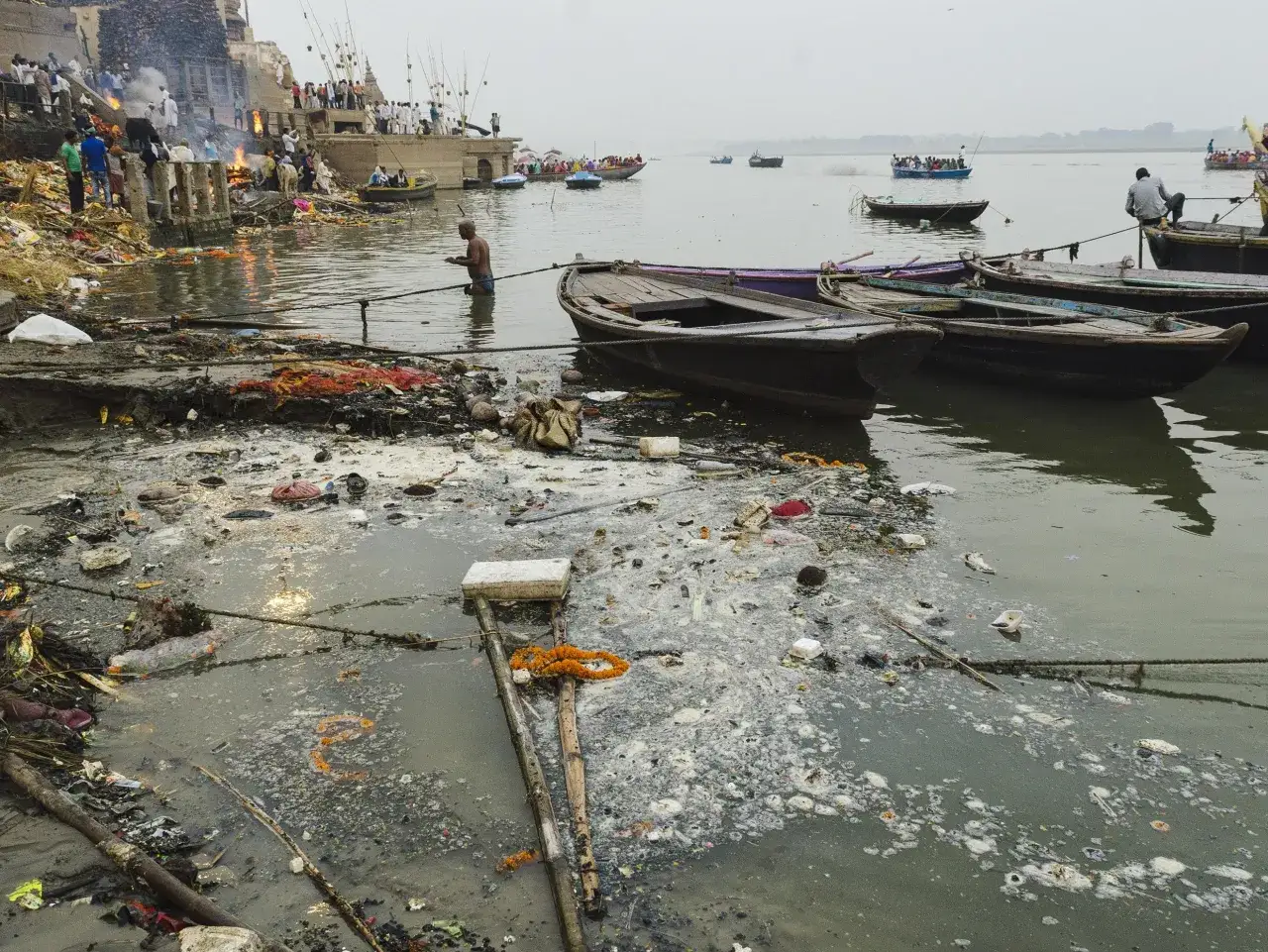 Rzeka Ganges, święta w hinduizmie, jest miejscem rytuałów i kremacji. Widać łodzie i ludzi na brzegu, ale też śmieci unoszące się na wodzie.