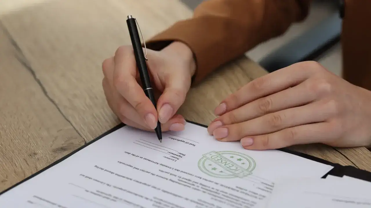 Dłoń podpisuje dokument, na którym widnieje pieczęć "LICENSED". To ważny krok do uzyskania licencji.