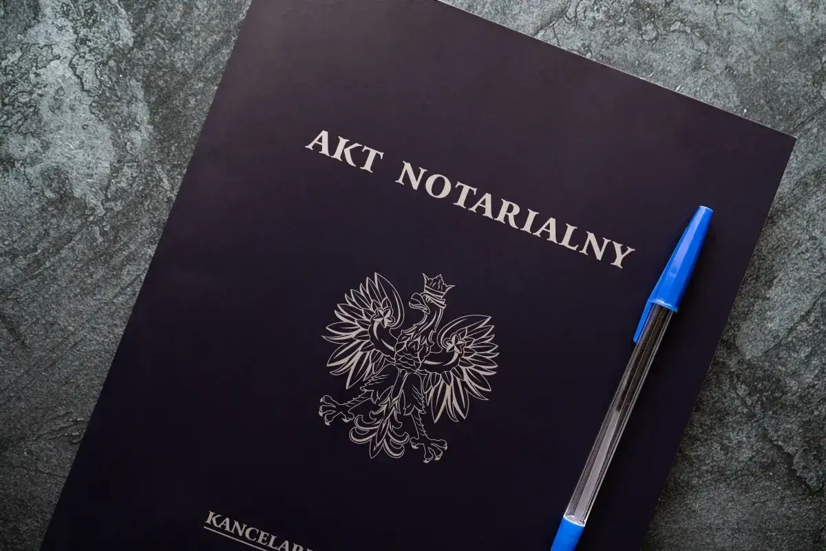 Na ciemnogranatowej okładce widnieje napis "AKT NOTARIALNY" i godło Polski. Obok leży niebieski długopis.