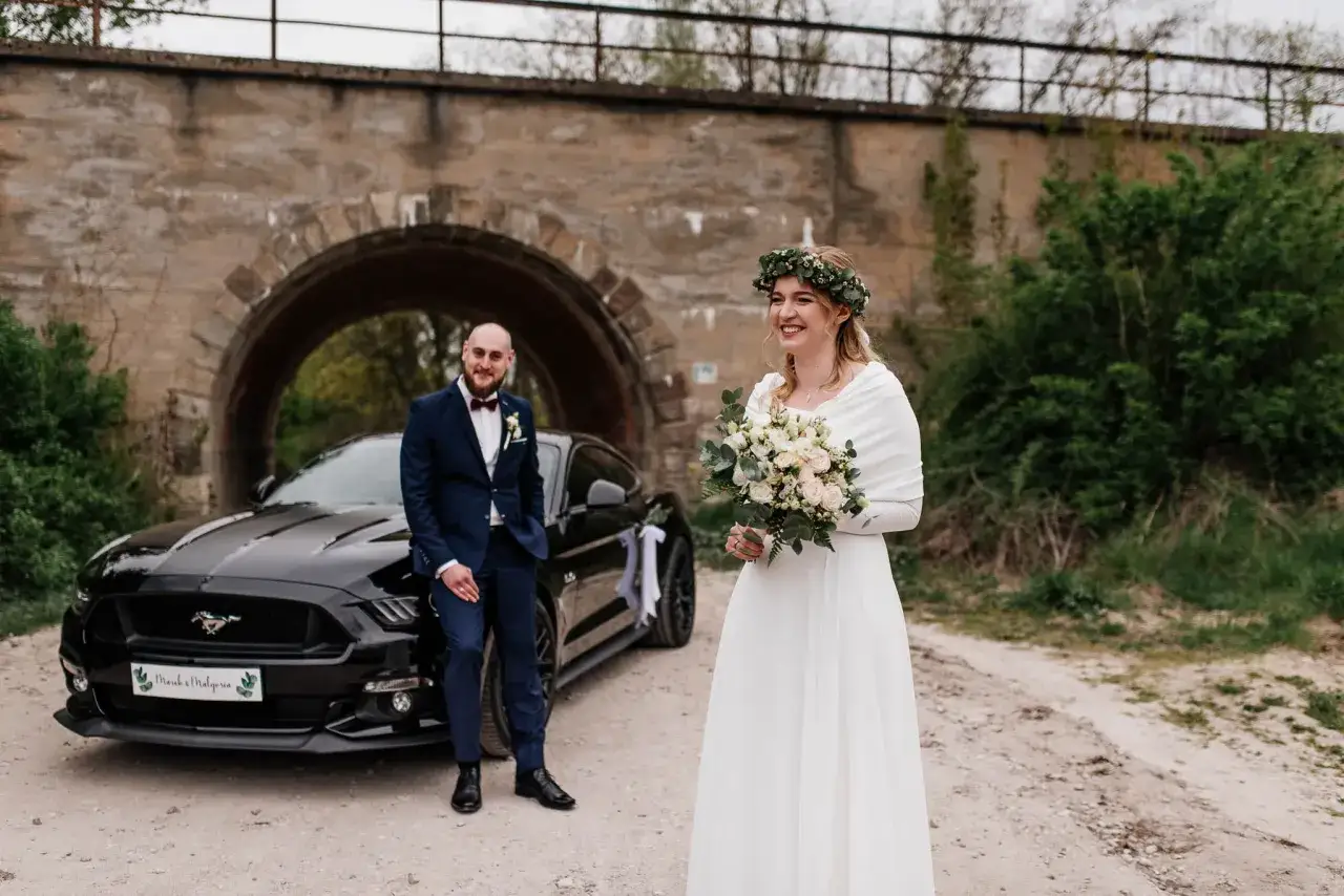 Para nietypowe sesje ślubne pozuje przy czarnym Mustangu pod kamiennym wiaduktem. Panna młoda w białej sukni i wianku z zieleni, pan młody w granatowym garniturze.