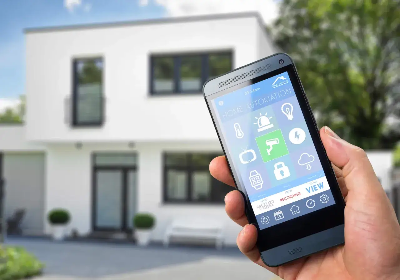 Dłoń trzyma smartfon z aplikacją do **instalacji inteligentnego domu**. Na ekranie widoczne ikony sterowania oświetleniem, ogrzewaniem, kamerami i innymi funkcjami.