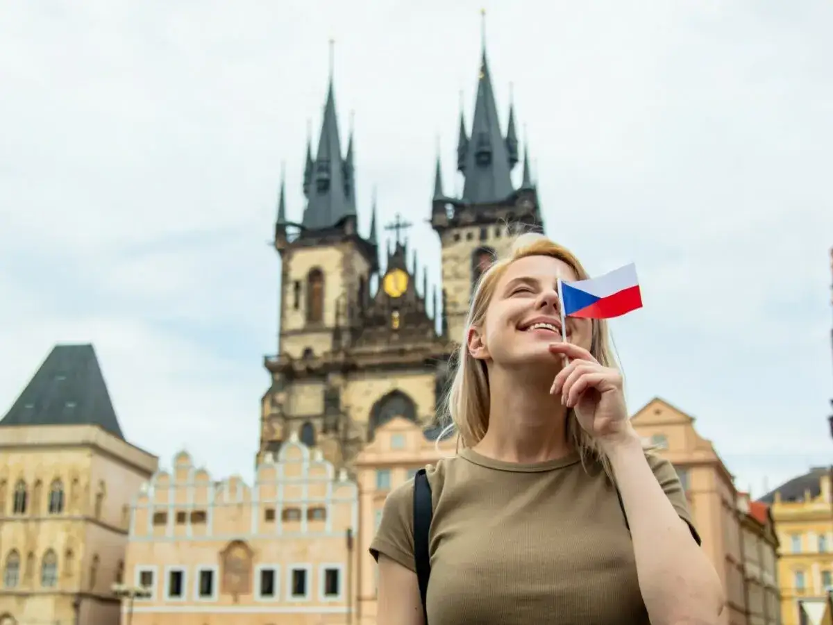 Uśmiechnięta blondynka z flagą Czech zasłaniającą oko, marzy o wysokich zarobkach w Czechach. W tle praskie kamienice i kościół.