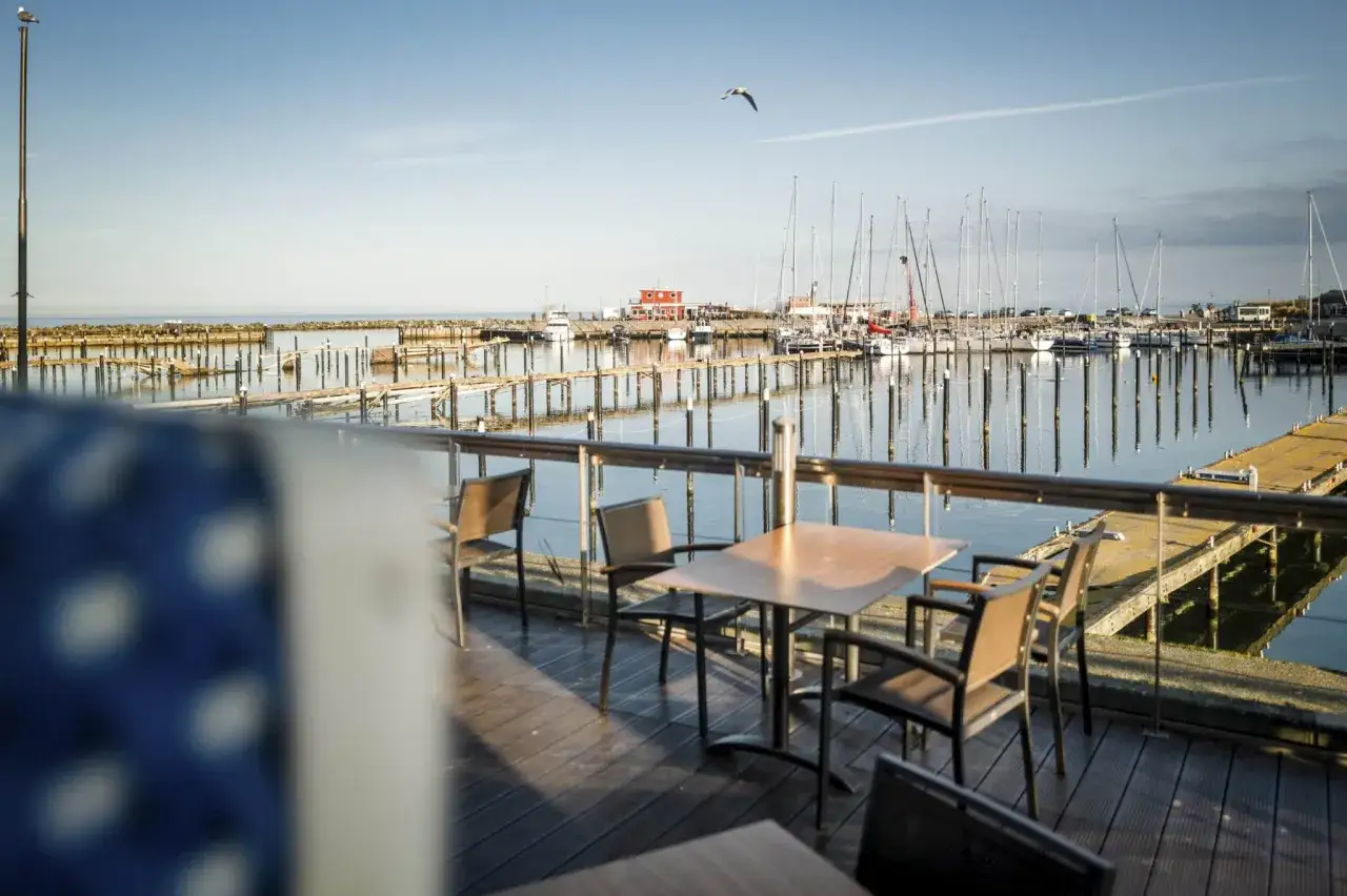 Ein Tisch mit Stühlen auf einer Holzterrasse mit Blick auf einen Yachthafen. Segelboote liegen im Wasser, die Sonne wirft lange Schatten. Ein Möwe fliegt über den Hafen. Damp 2000.