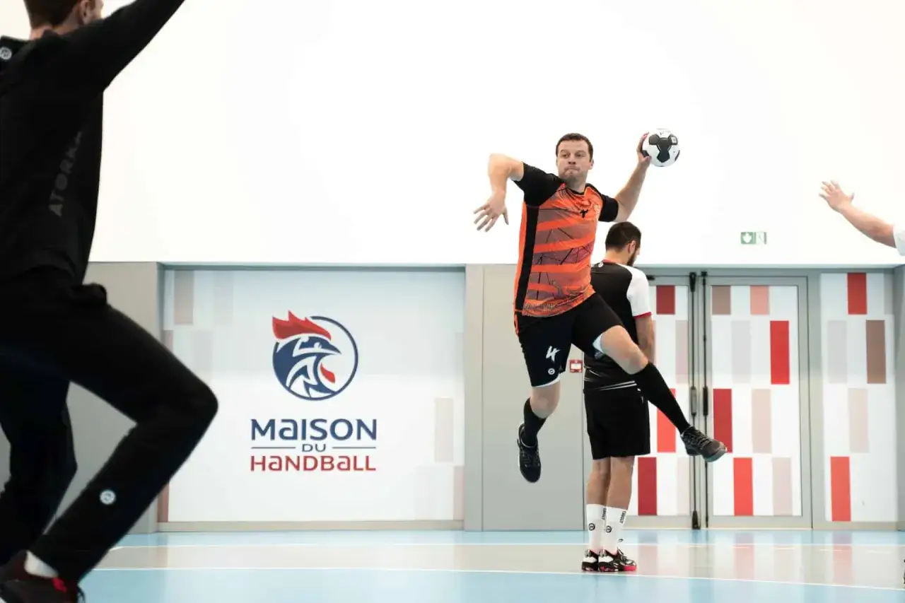 Piłkarz ręczny, niczym szczypiornista, wyskakuje do rzutu. W tle logo "Maison du Handball".