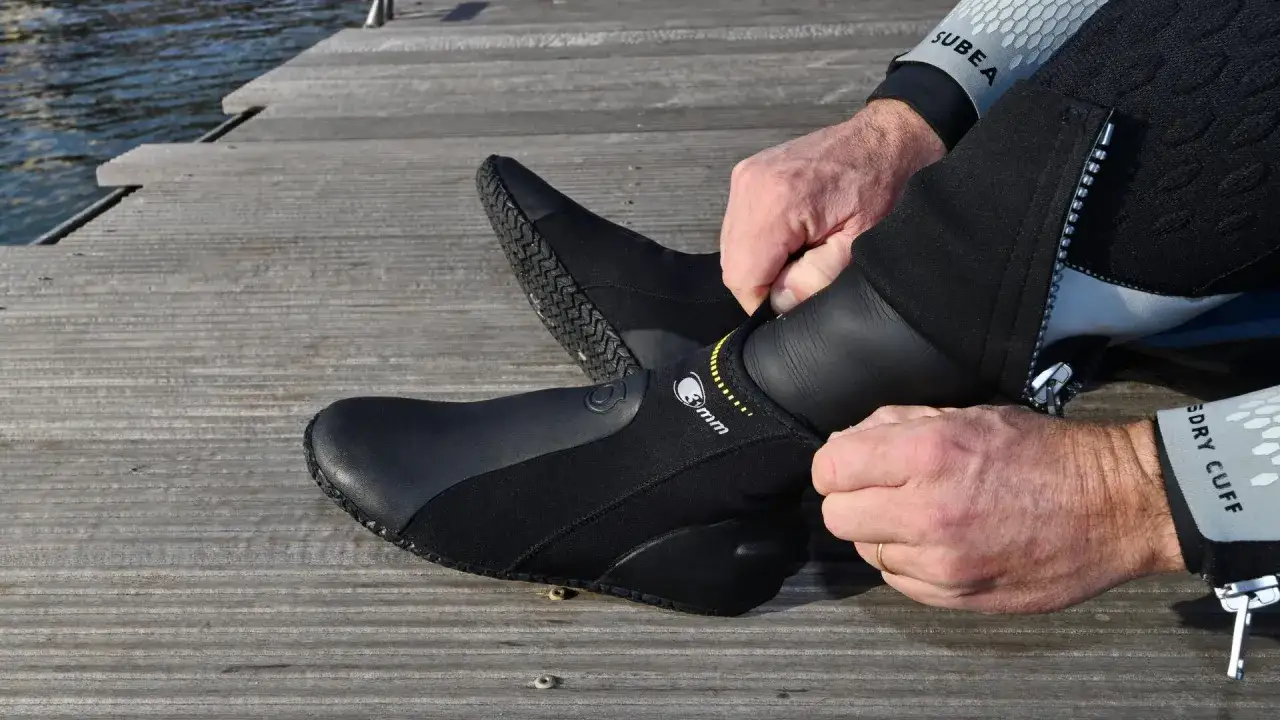 Mężczyzna zakłada czarne buty neoprenowe na drewnianym pomoście przy wodzie.