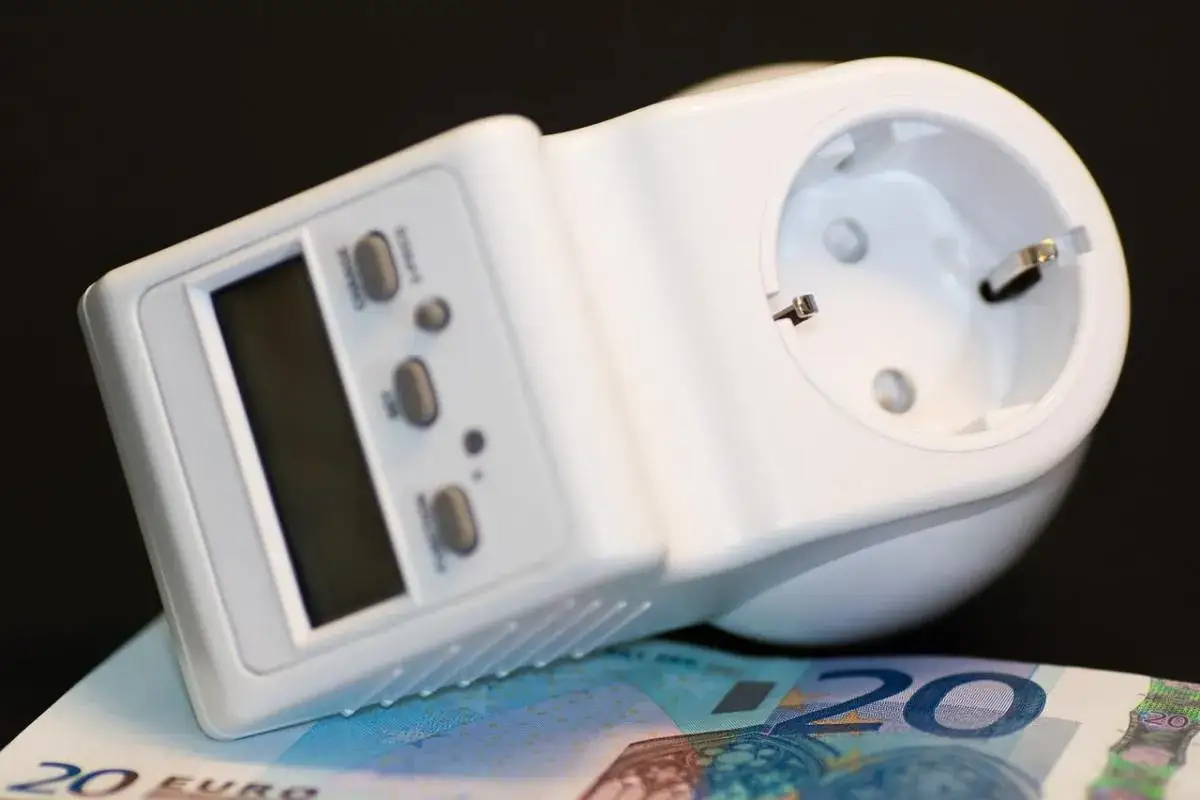 Biały miernik zużycia prądu z wyświetlaczem i przyciskami, leżący na banknocie 20 euro.