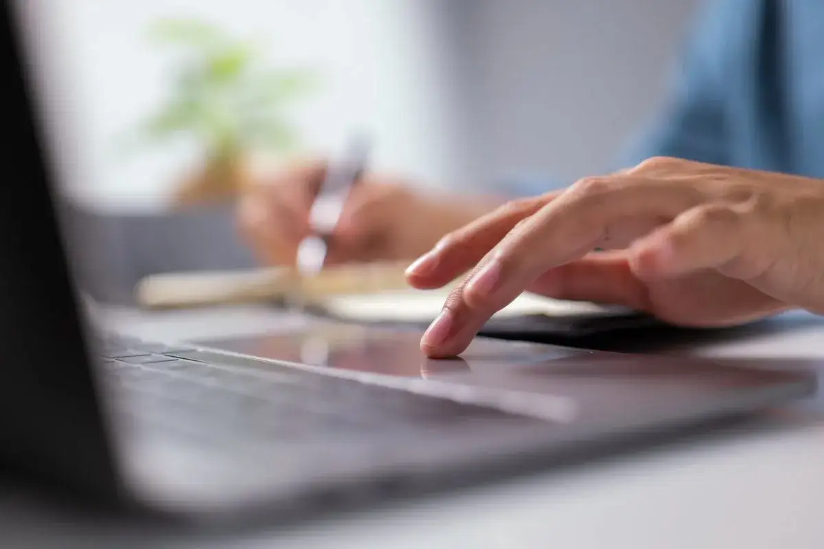 Dłoń na touchpadzie laptopa, w tle notatnik i długopis.