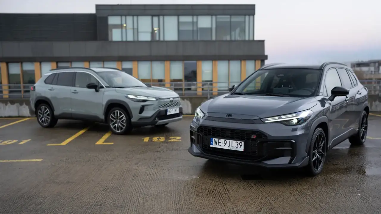 Dwie nowe Toyoty Corolla Cross, jedna szara, druga ciemnoszara GR, zaparkowane na mokrym parkingu.
