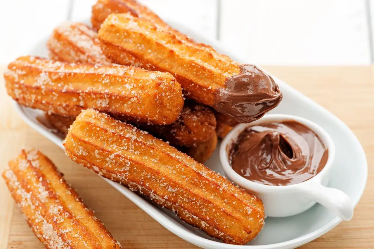 Churros posypane cukrem, jeden zanurzony w czekoladzie, obok miseczki z kremem czekoladowym.