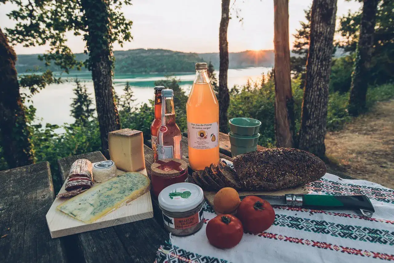 Pique-nique avec fromages, pain de campagne, jus de poire et terrine, un aperçu des spécialités culinaires Jura au bord d'un lac.