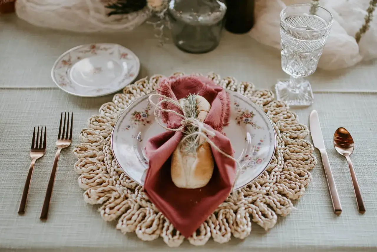 Une table dressée pour un repas, avec une assiette fleurie, une serviette bordeaux, un petit pain et une brindille de romarin. L'esprit **dressage à la française** est palpable.
