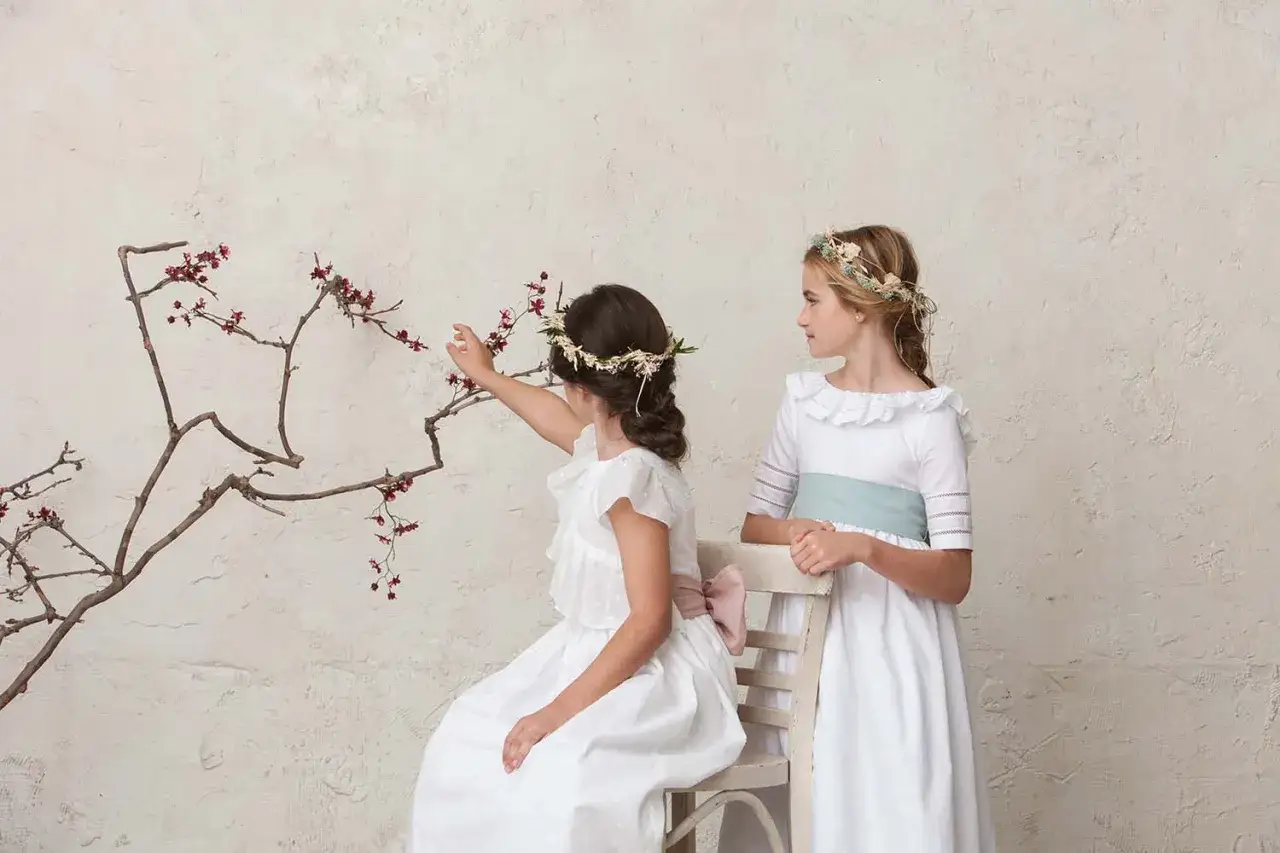 Dos niñas con vestidos blancos y coronas de flores, una sentada tocando una rama, la otra de pie. Peinados de trenza para niña de comunión.