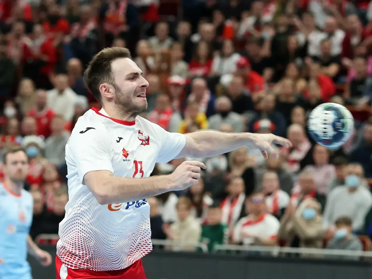 Piłkarz ręczny Polski w akcji, rzuca piłkę. W tle widać trybuny z kibicami, którzy dopingują swoją drużynę w meczu przeciwko Iranowi.