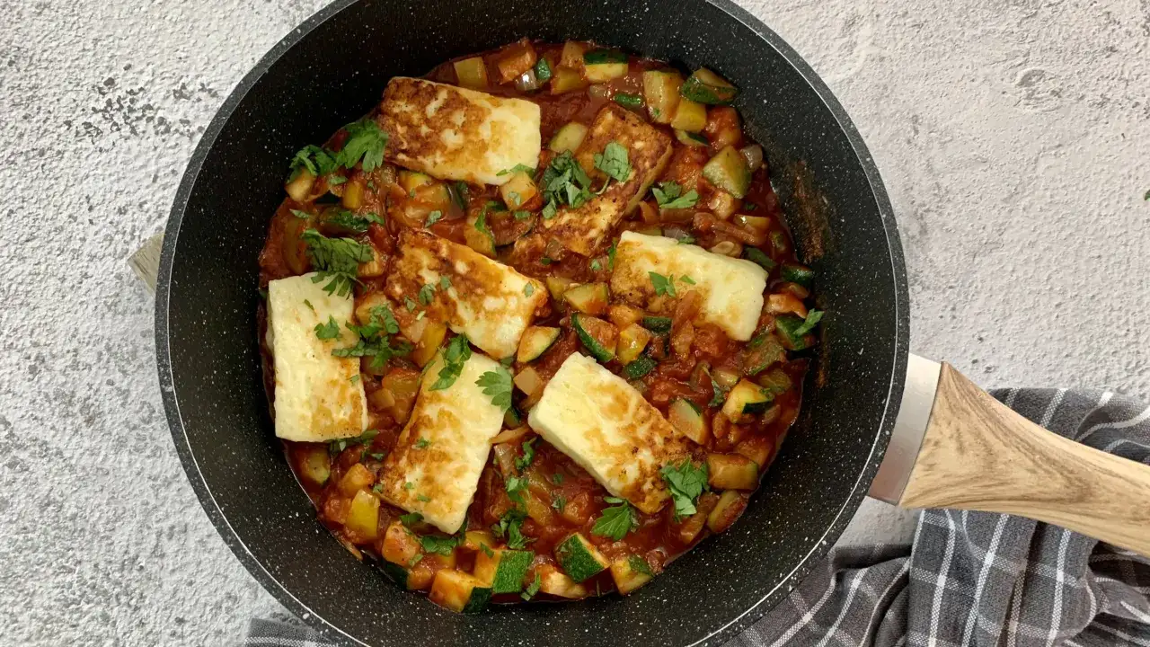 Smażony ser halloumi z warzywami w sosie pomidorowym, posypany natką pietruszki, w czarnej patelni.