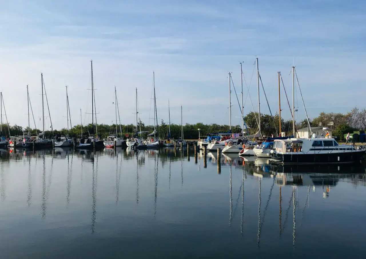 Segelboote und Motorboote liegen friedlich im Hafen Neuendorf, ihre Spiegelbilder tanzen auf dem ruhigen Wasser.