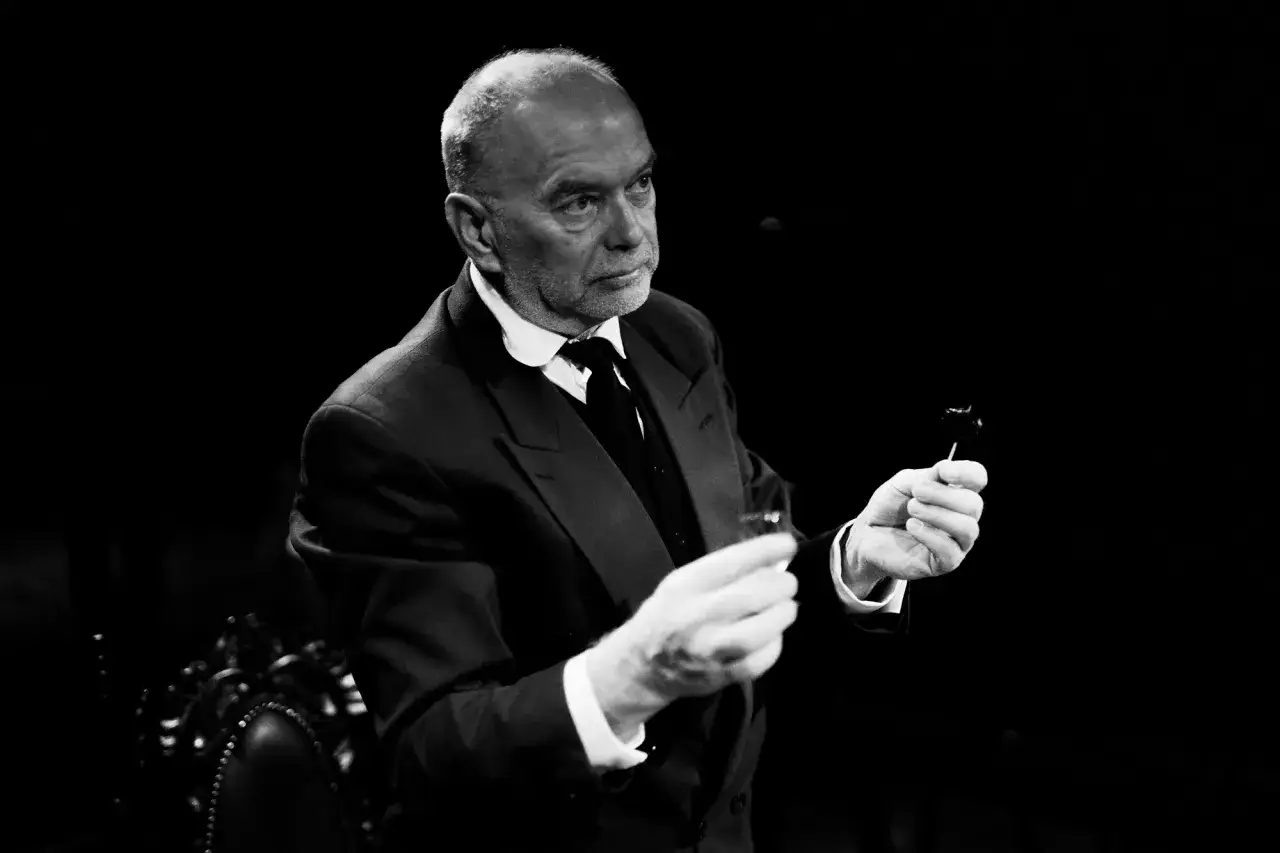 Piotr Wyszomirski w eleganckim garniturze, trzymający w dłoniach kieliszek i coś na patyczku.