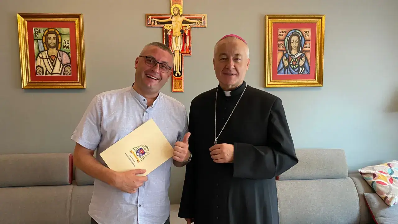 Uśmiechnięty mężczyzna z dokumentem i biskup pozują na tle krzyża i obrazów. Mężczyzna trzyma dokument z herbem, a biskup ma na sobie sutannę.