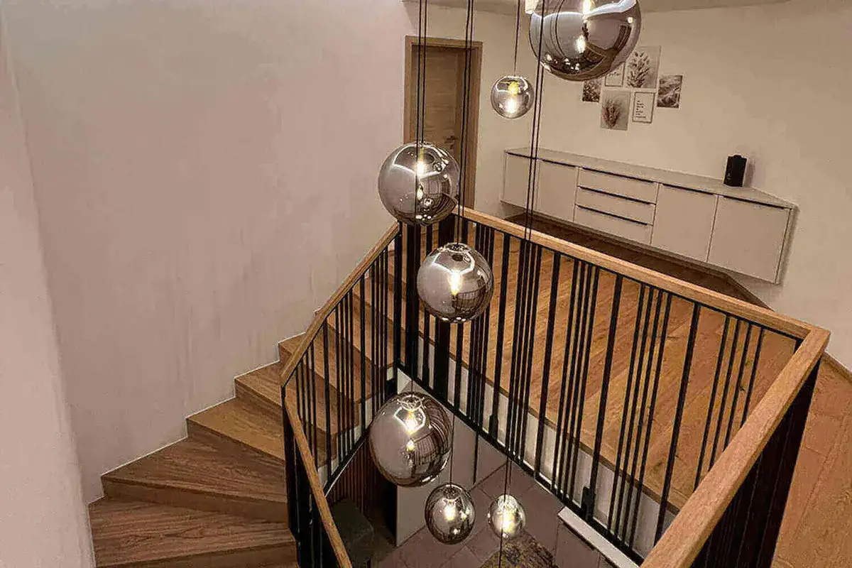 Nowoczesna klatka schodowa w domu z drewnianymi stopniami i czarną balustradą, ozdobiona wiszącymi, lustrzanymi kulami.