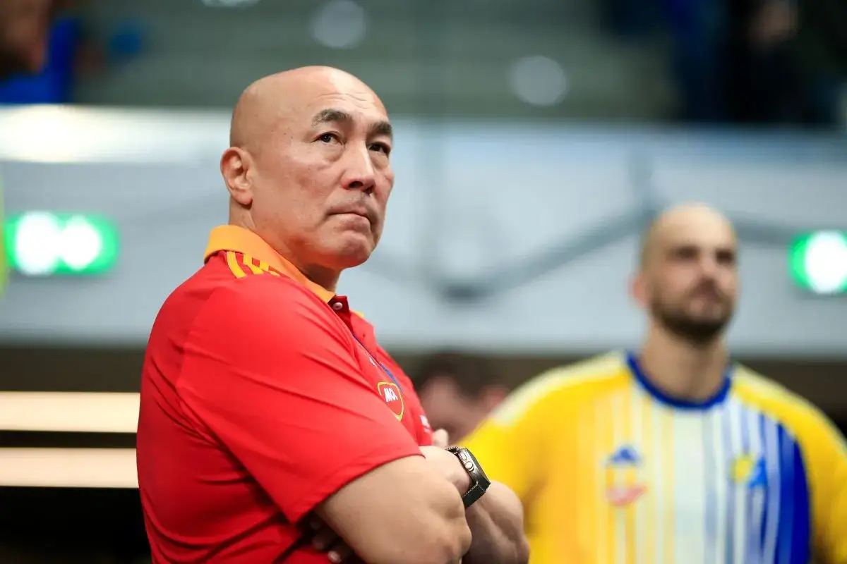 Skupiony trener Vive Kielce w czerwonej koszulce z pomarańczowym kołnierzem, z rękami skrzyżowanymi na piersi.