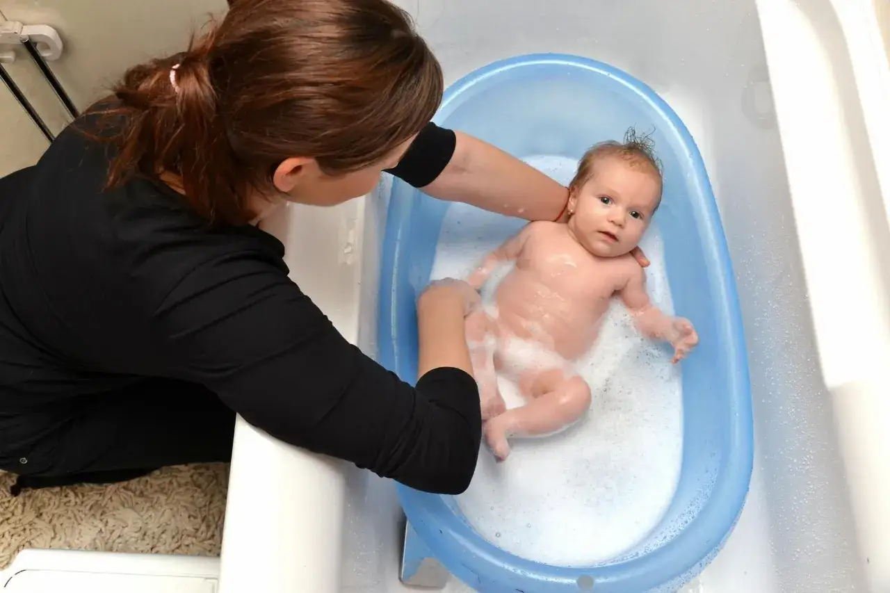 Mama delikatnie myje noworodka w niebieskiej wanience. Maluch pluska się w pianie, a mama uśmiecha się do niego.