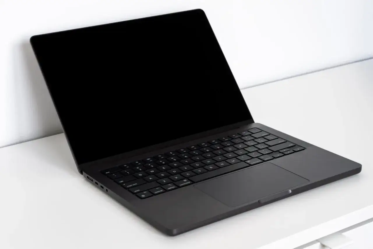 Ciemny laptop z czarnym ekranem, wyglądający jak Mac OS na PC, stoi na białym biurku.