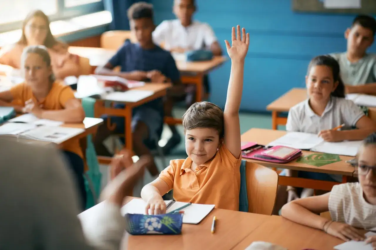 Chłopiec z podniesioną ręką czeka na ocenianie odpowiedzi. Inne dzieci pracują przy ławkach.
