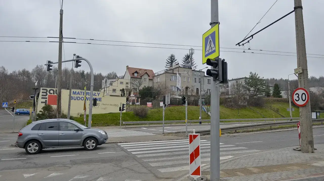 Szare auto zbliża się do przejścia dla pieszych, gdzie światła kolizyjne regulują ruch. W tle budynki i drzewa.