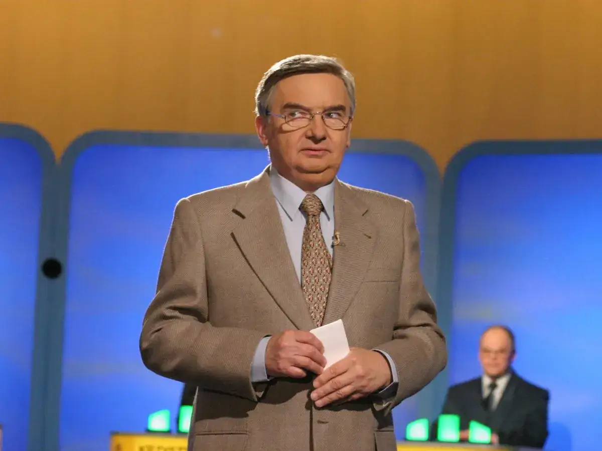 Tadeusz Sznuk, który prowadzi teleturniej Jeden z dziesięciu, w beżowej marynarce i z kartką w dłoni.