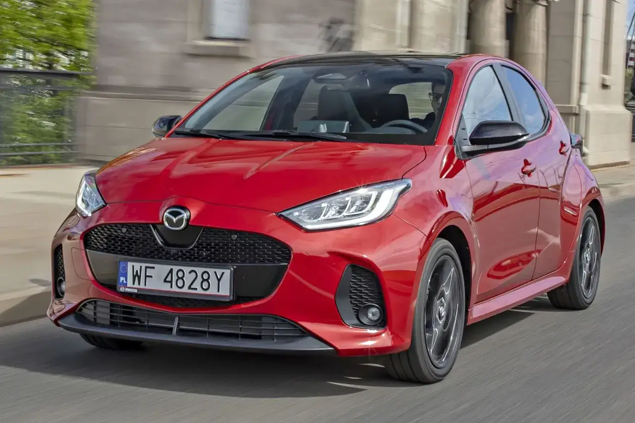 Czerwona Mazda 2 Hybrid w ruchu miejskim, z widocznym grillem i reflektorami.