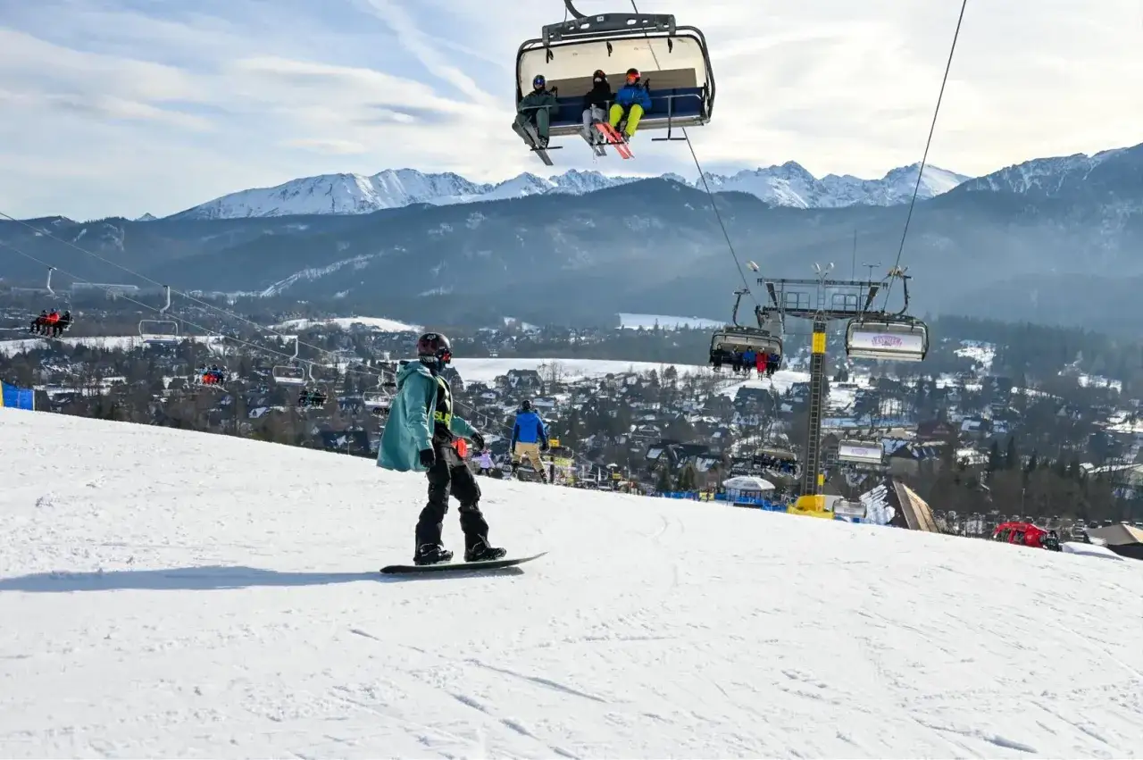 Snowboardzista na stoku, w tle kolejka krzesełkowa i góry. Zastanawiasz się, ile kosztuje karnet na narty?