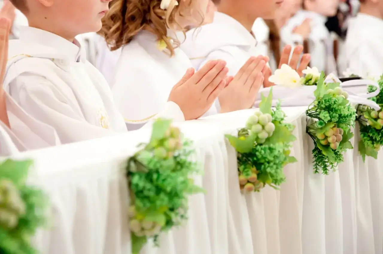 Dzieci w bieli podczas komunii, modlące się ręce. Dekoracje stołu z winogronami i zielenią, symbolizujące koszty komunii w kościele.
