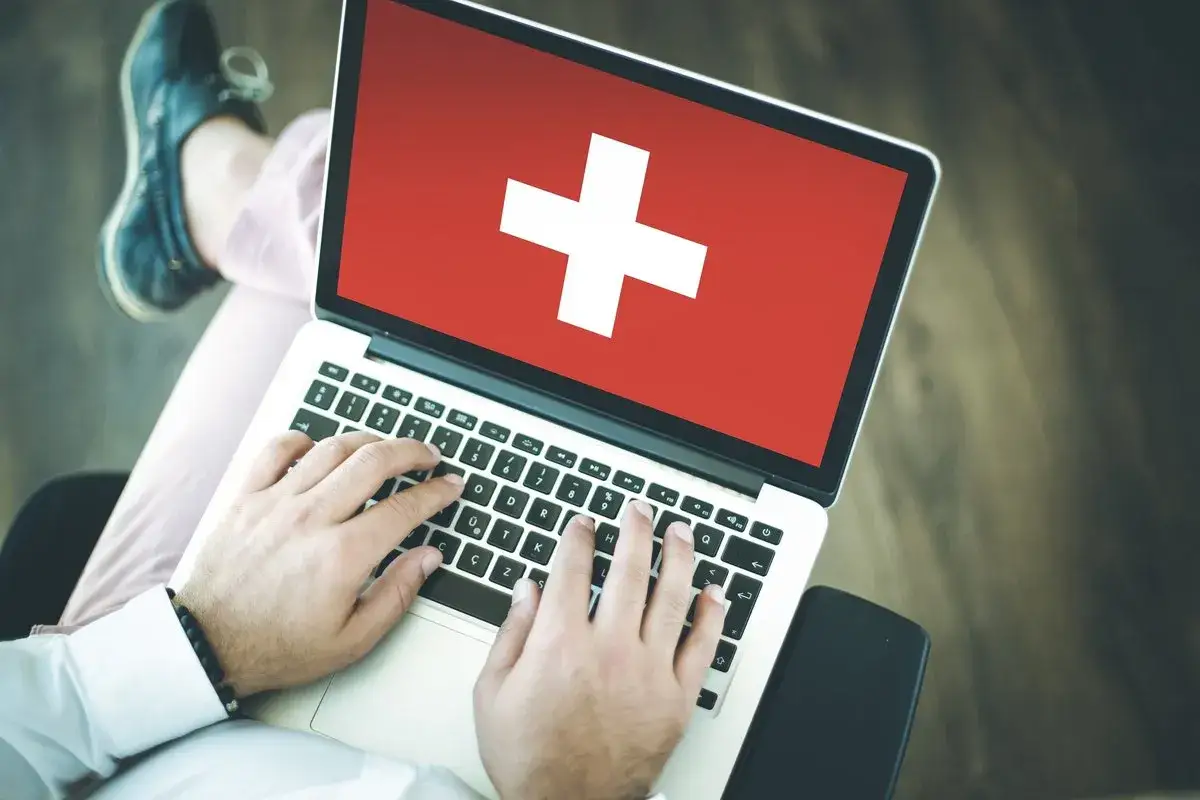 Osoba pisząca na laptopie z flagą Szwajcarii na ekranie.