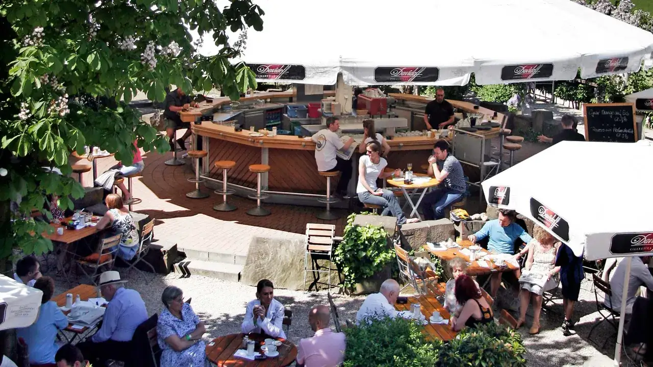 Gäste sitzen an Tischen und an der Theke eines Biergartens unter Sonnenschirmen.