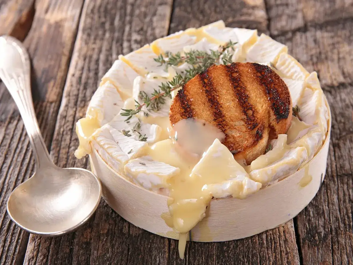 Un camembert coulant, plat typique normandie, garni d'une tranche de pain grillé et de thym, prêt à être dégusté avec une cuillère.