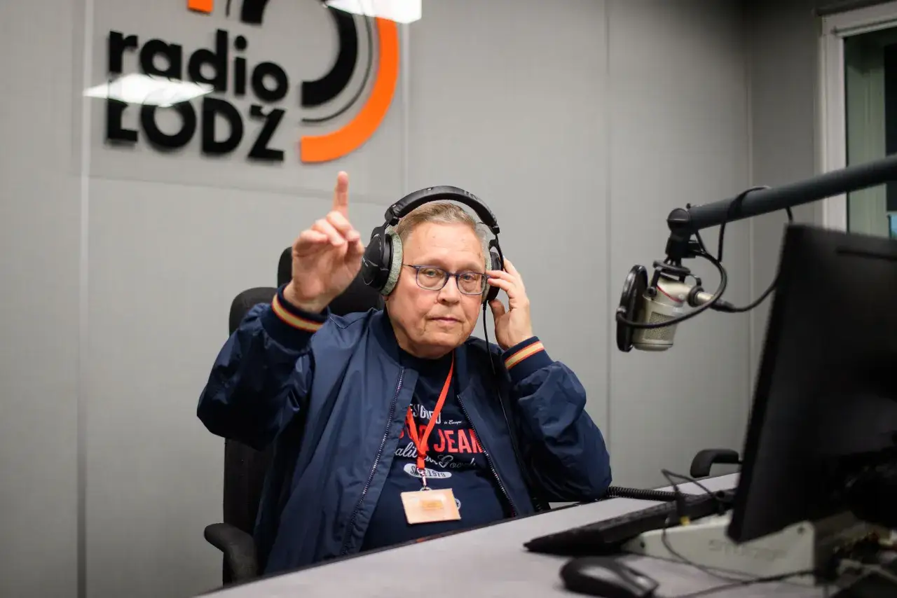 Mężczyzna w słuchawkach i okularach, siedzący przy mikrofonie w studiu radiowym. Wskazuje palcem, jakby coś tłumaczył. Ale numer obsada!