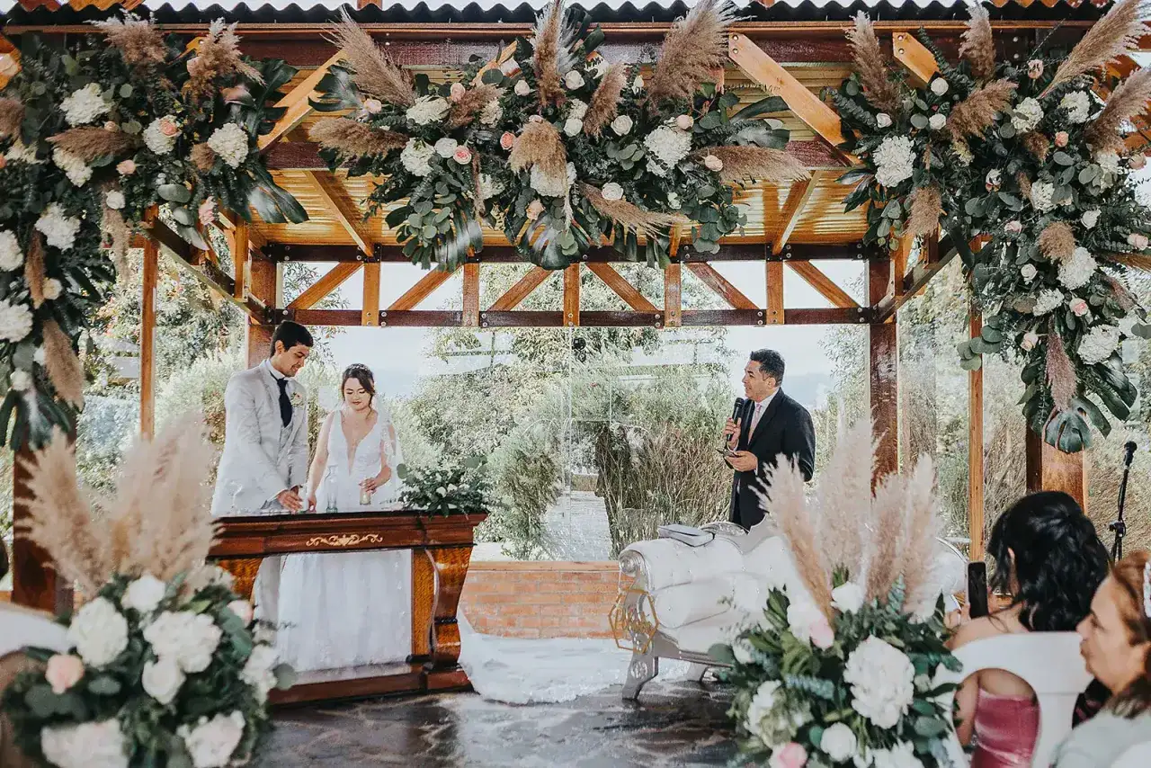Novios celebran aniversario en altar decorado con flores y pampas. Un oficiante habla mientras la pareja sonríe.