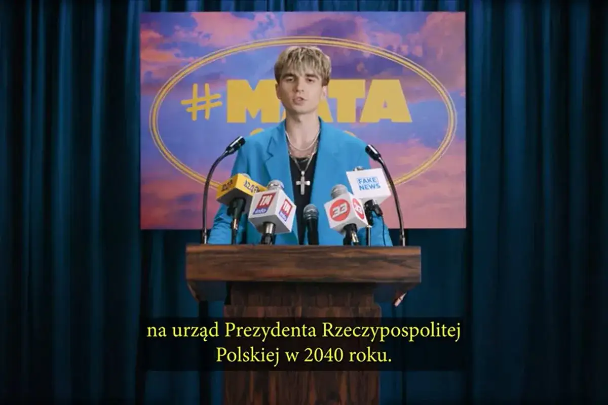 Młody mężczyzna w niebieskiej marynarce przemawia za mównicą z mikrofonami.