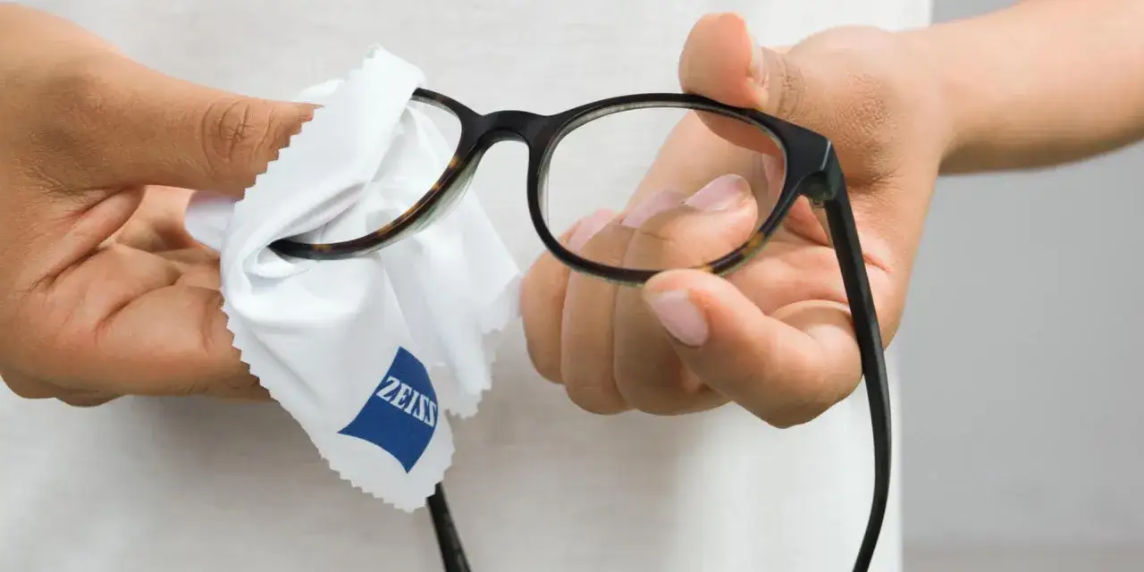 Dłonie czyszczą okulary okularowe białą ściereczką z logo Zeiss.