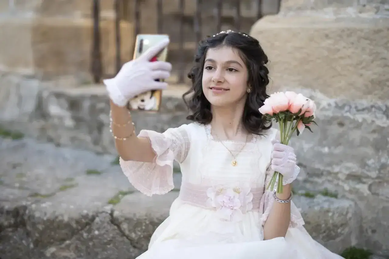 Dziewczynka w sukni komunijnej robi selfie z bukietem kwiatów. To ważny moment, przygotowania do pierwszej komunii świętej krok po kroku.