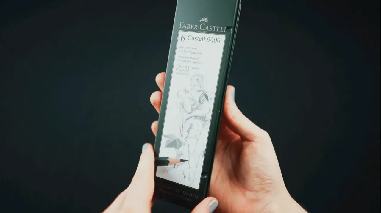Dłonie trzymają opakowanie z sześcioma ołówkami Faber-Castell 9000. Jeden ołówek rysuje szkic postaci.
