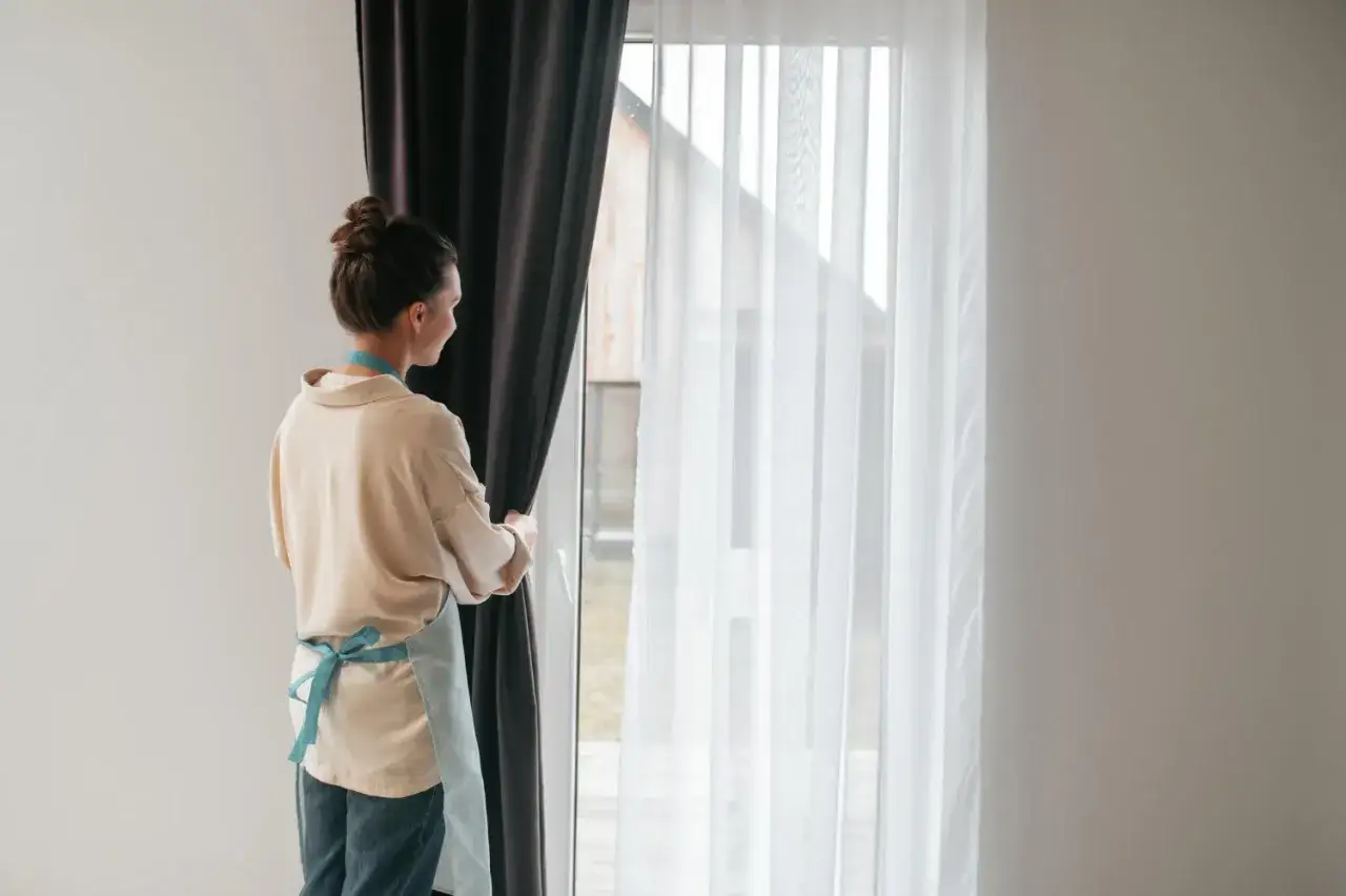 Kobieta w fartuszku zasłania okno ciemną zasłoną. Widać białe firanki i karnisz z wieloma ząbkami.