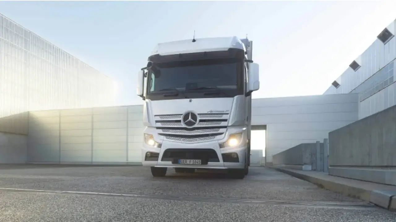 Biały Mercedes Actros MP4 z włączonymi światłami stoi przed nowoczesnym budynkiem.