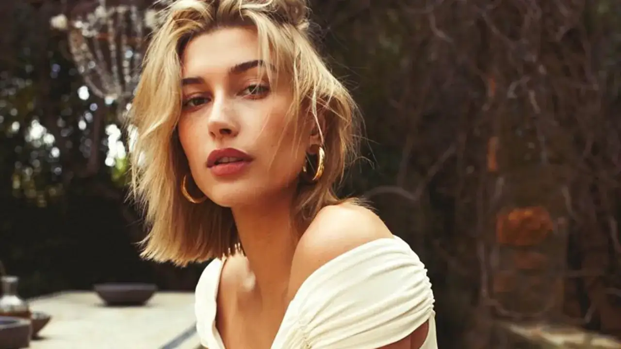 Mujer con un moderno corte bob rubio, aretes dorados y un vestido blanco con hombros descubiertos, posando al aire libre.