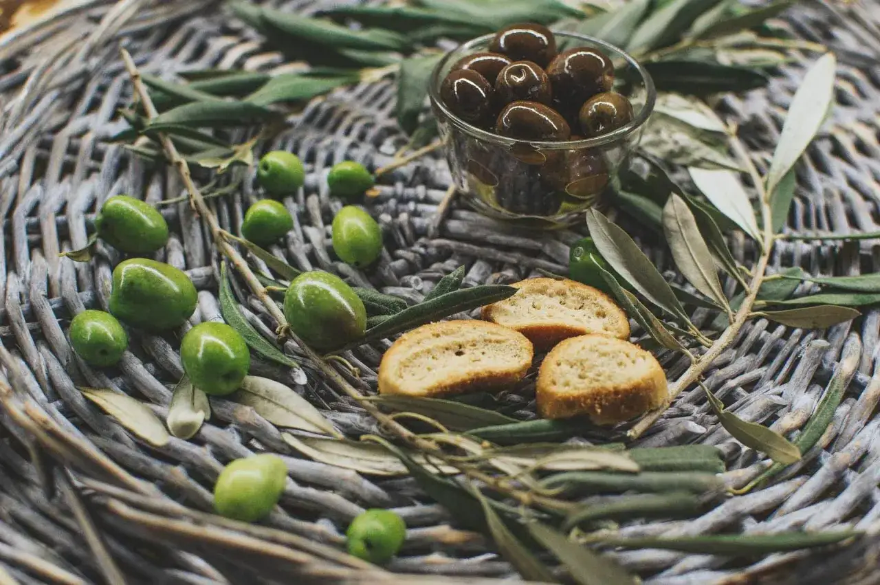 Olives vertes et noires, crostinis et feuilles d'olivier sur un panier tressé, évoquant une spécialité culinaire du Luberon.