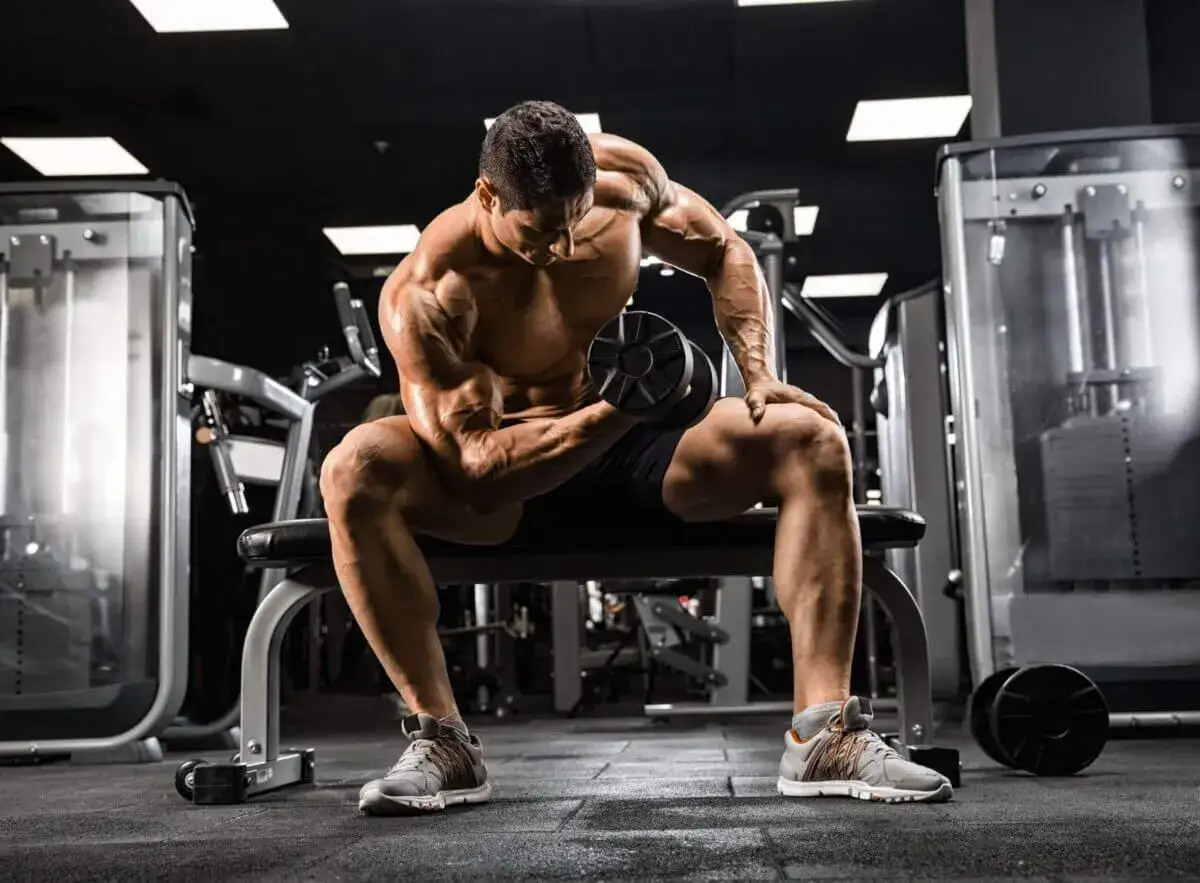 Mężczyzna z wyrzeźbionymi mięśniami wykonuje ćwiczenie na biceps z hantlem, skupiony na swoim **full body workout** w siłowni.