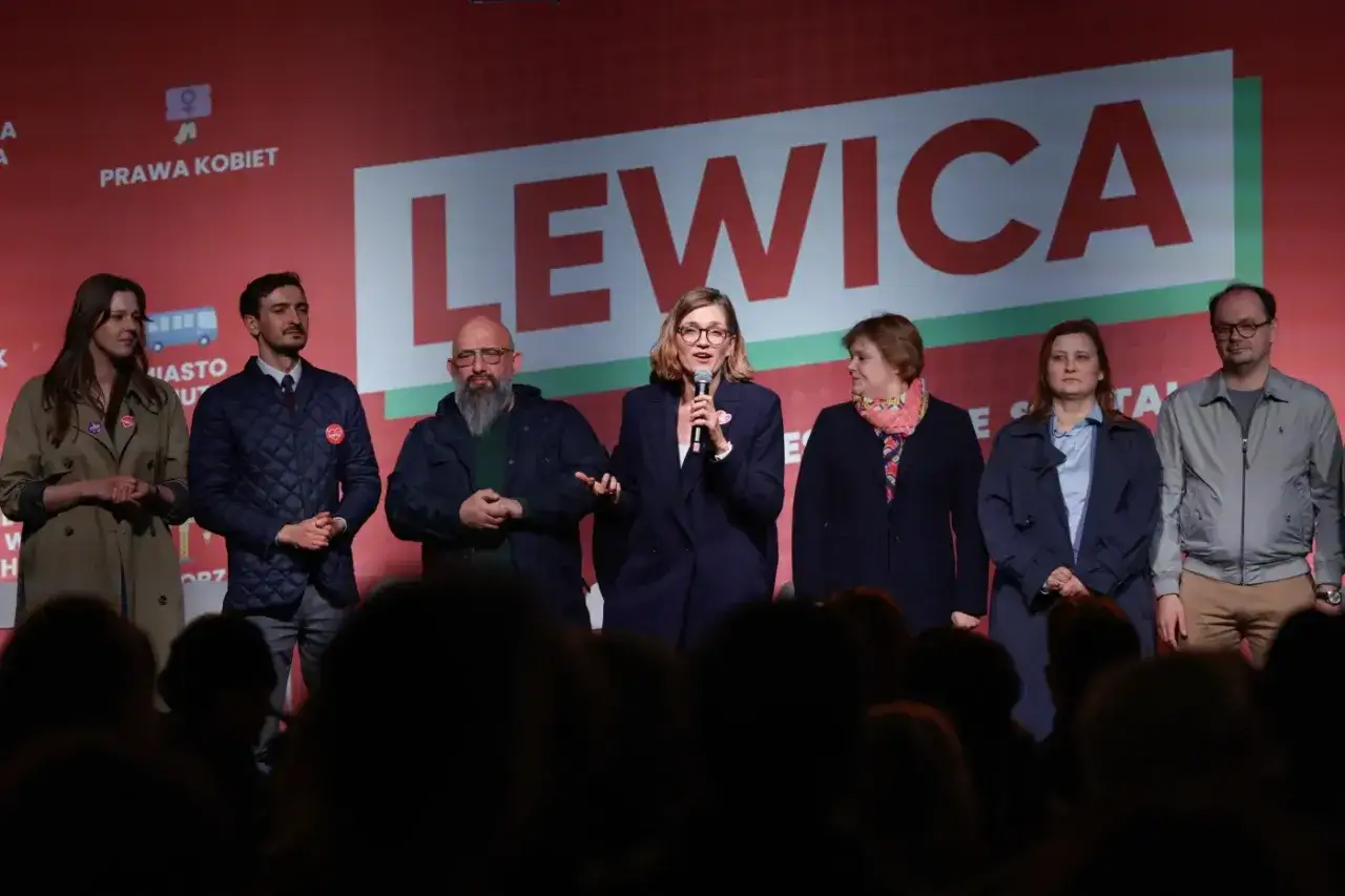 Grupa osób stoi na scenie przed napisem "LEWICA". Kobieta w środku mówi do mikrofonu.